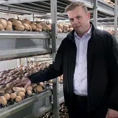 Mężczyzna w czarnej kurtce stoi obok regałów pełnych pieczarek w hali produkcyjnej.