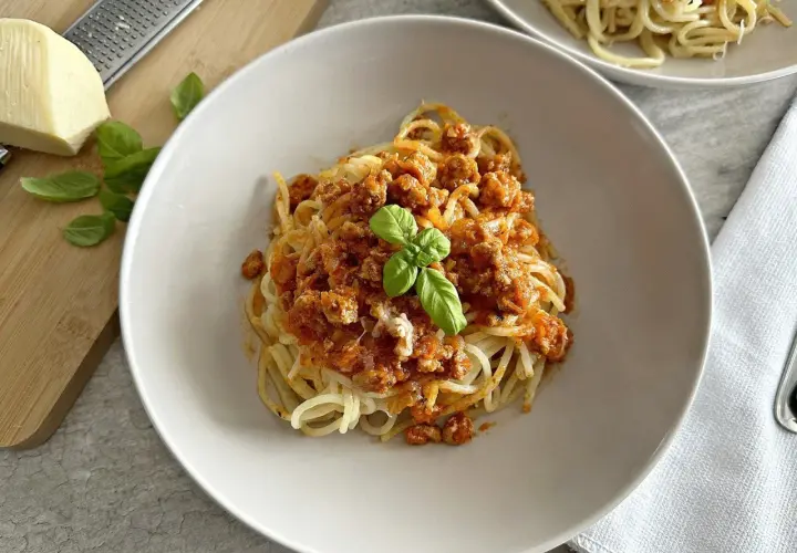 Spaghetti z sosem mięsnym, udekorowane bazylią, na talerzu. Obok ser i tarka.