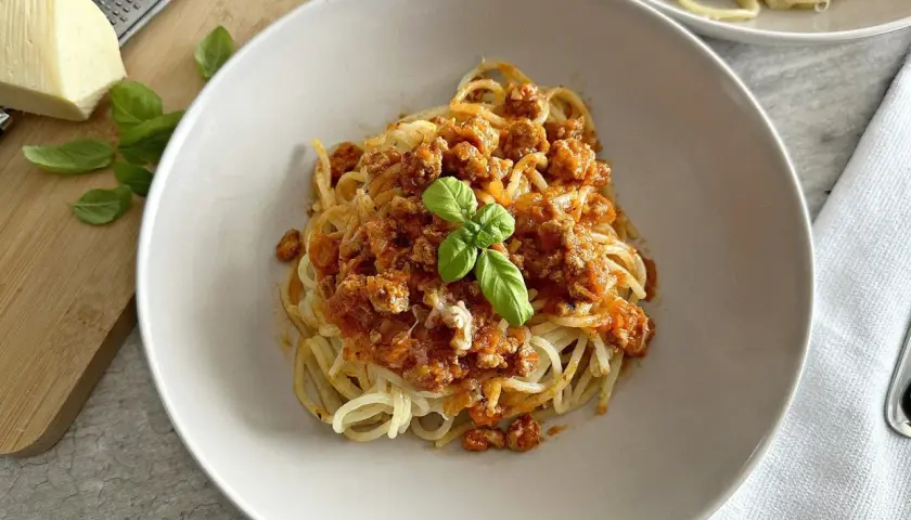 Spaghetti z sosem mięsnym, udekorowane bazylią, na talerzu. Obok ser i tarka.