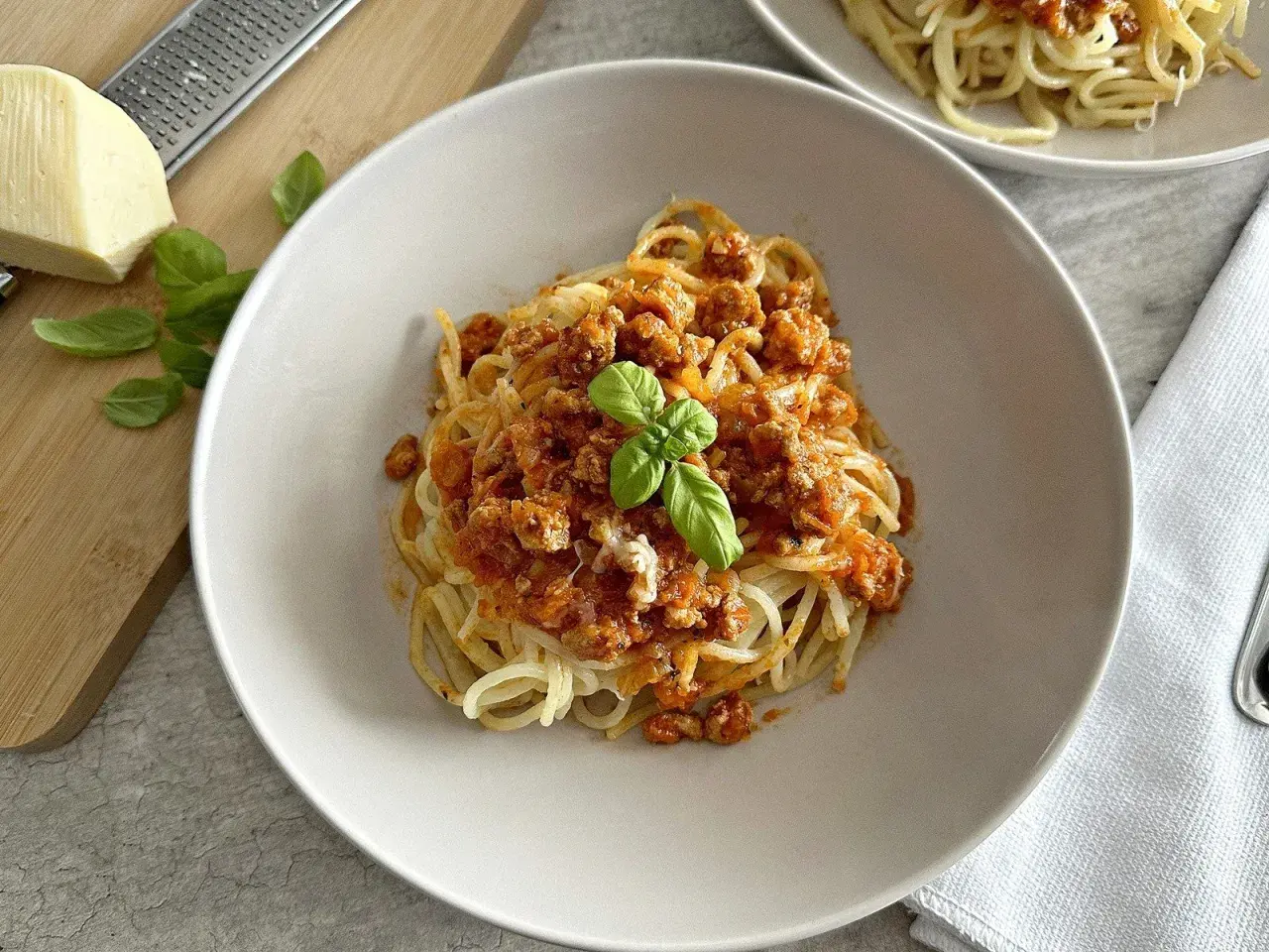 Spaghetti z sosem mięsnym, udekorowane bazylią, na talerzu. Obok ser i tarka.